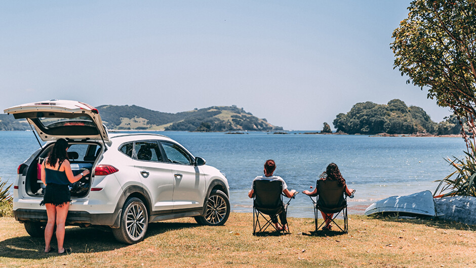 Group of friends sitting in lawn chairs by the water with a GO Rentals car.