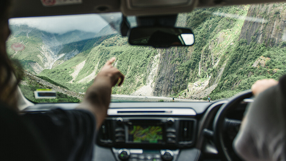 Two friends sitting in a GO Rentals car looking out the windscreen pointing at the green scenery.